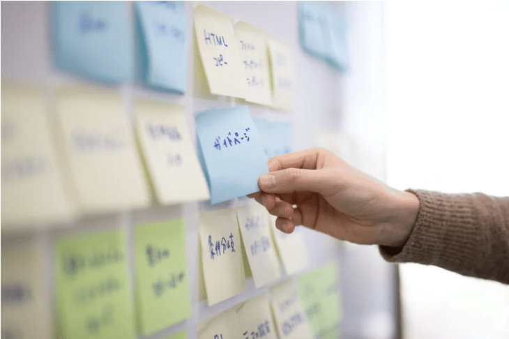 Una pared de post its, algunos con palabras como "html", otros en japonés, y una mano cogiendo uno de ellos.