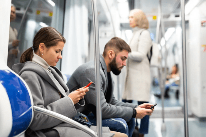 Dos personas sentadas en el metro mirando el móvil, y en segundo plano una persona de pie mirando la puerta.